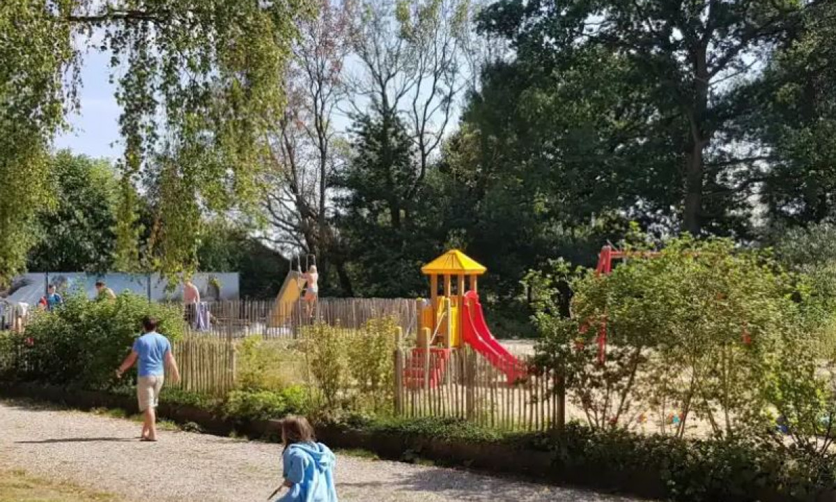 At Elbeling, kids are playing on play structures encircled by trees and shrubs, with a pathway in the foreground where more children are present.