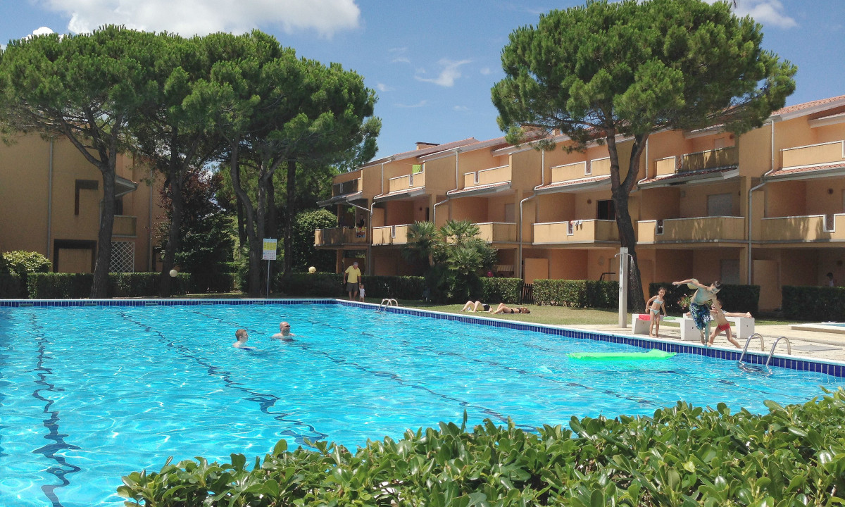 Swimming pool with people swimming and playing, surrounded by trees and beige apartment buildings at Villaggio Selene