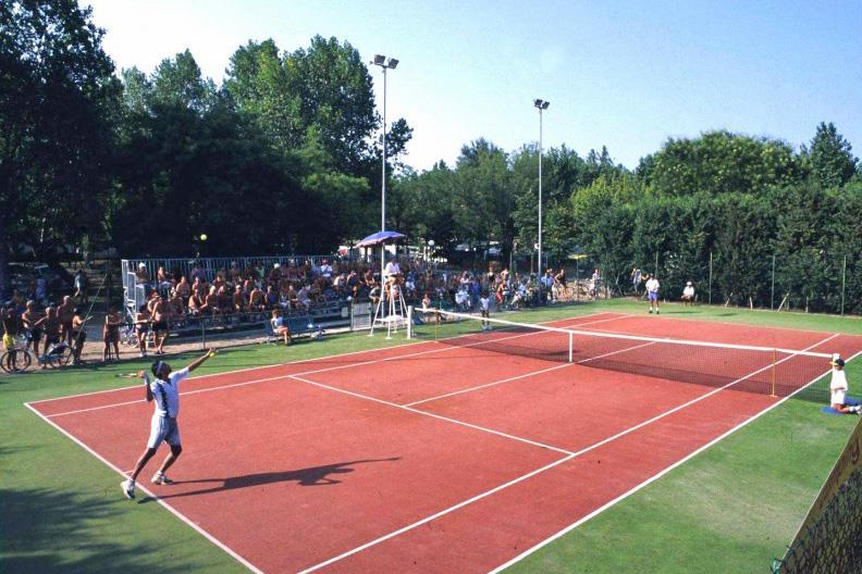 Tennis players are mid-game on a red court with a large audience seated on bleachers in a wooded area at Pineta Sul Mare Camping Village