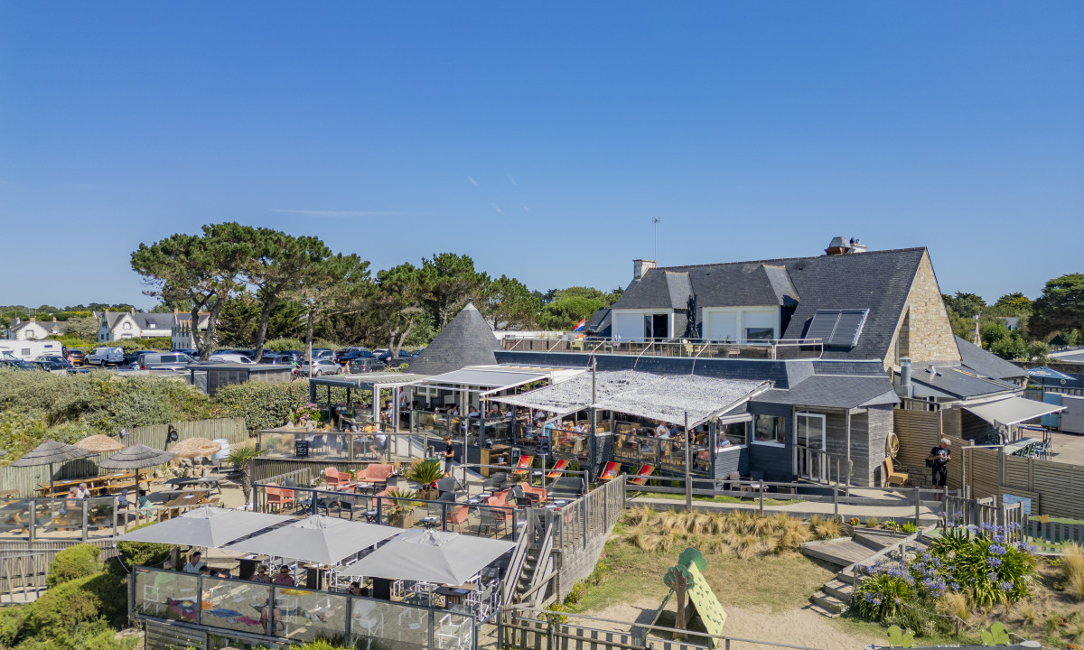 At De la Plage, a lively outdoor café is filled with patrons, encircled by lush vegetation and parked vehicles, all under a bright blue sky.