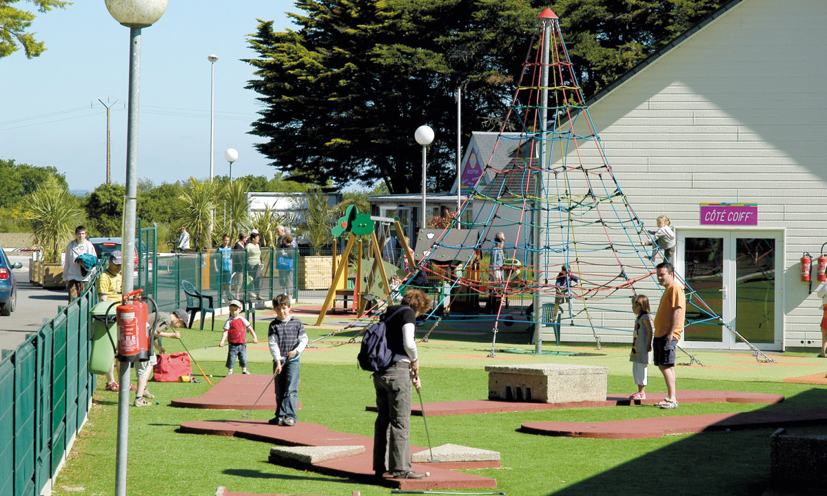 At Le Domaine de Léveno, individuals are engaging in mini-golf and navigating climbing play structures in an outdoor playground space adjacent to a building, with trees in the backdrop.