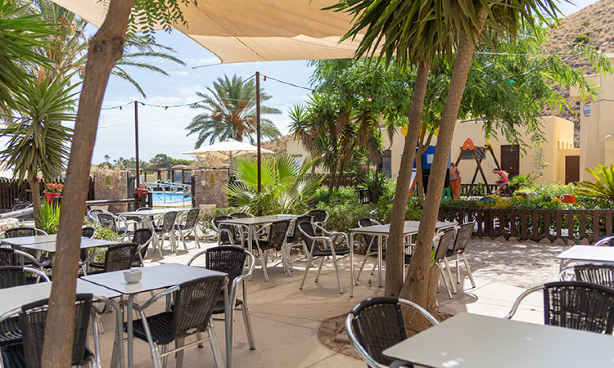 At Camping Los Escullos, metal seating and tables are positioned beneath a sunshade, encircled by palm trees and lush vegetation.