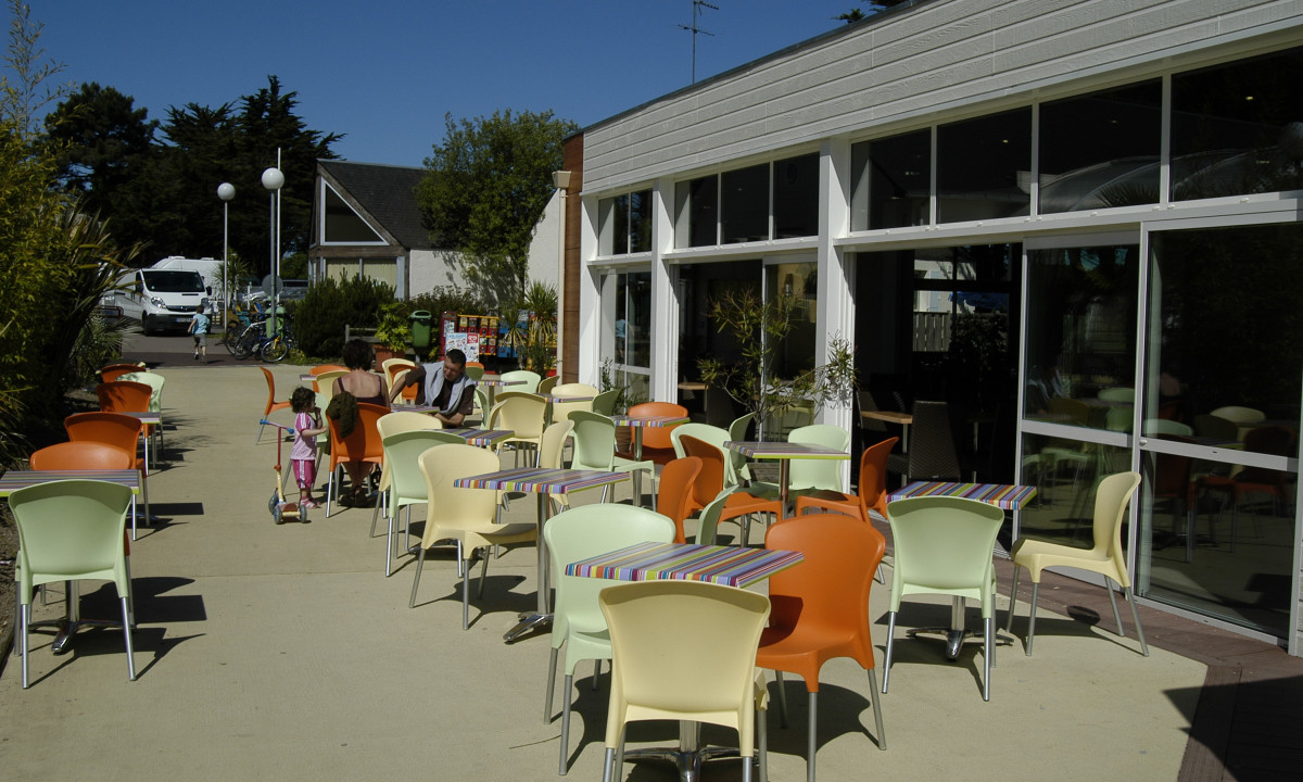 At Le Domaine de Léveno, vibrant chairs and tables are positioned outside a structure, with individuals seated and children strolling in a sunny outdoor space.