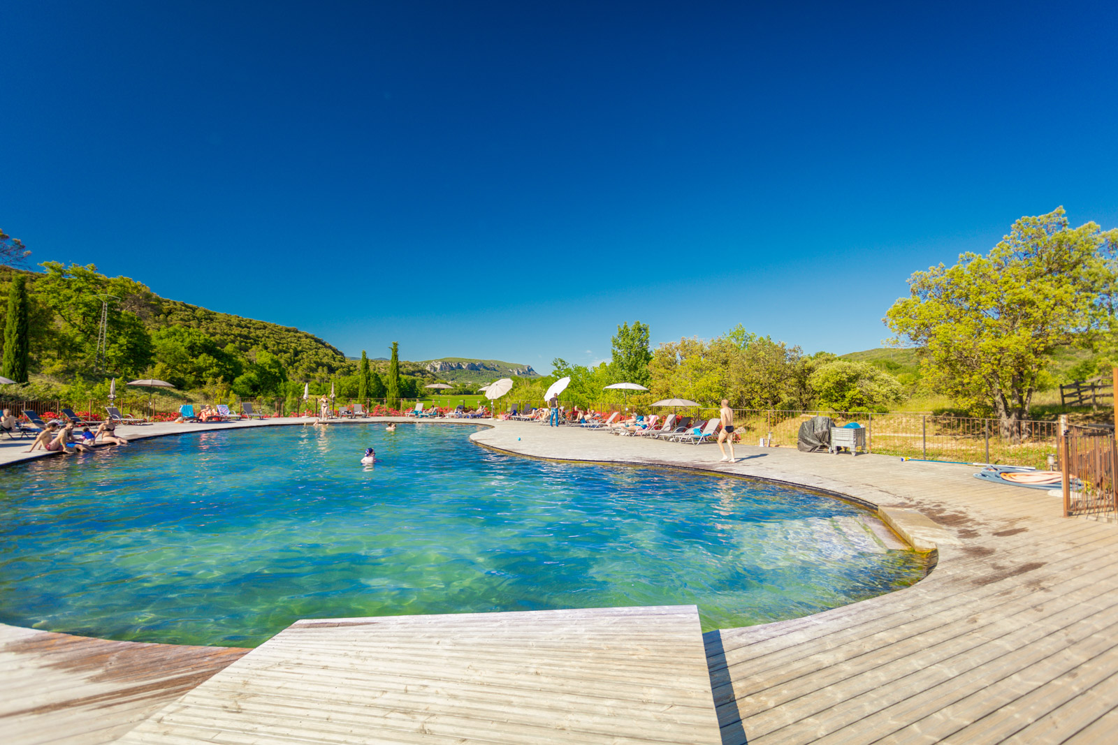 Swimming pool with people lounging and swimming, surrounded by trees and hills under a clear blue sky at du Domaine d'Anglas
