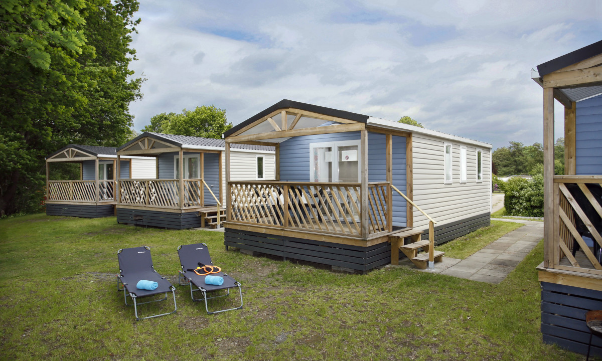 At KNAUS Campingpark Oyten, there are multiple cozy cabins featuring porches and two reclining chairs positioned in front on a grassy area.