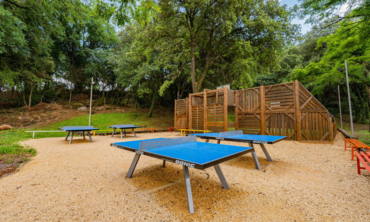 At La Clémentine, ping pong tables are positioned on gravel in a forested outdoor recreational space, encircled by trees and a wooden barrier.