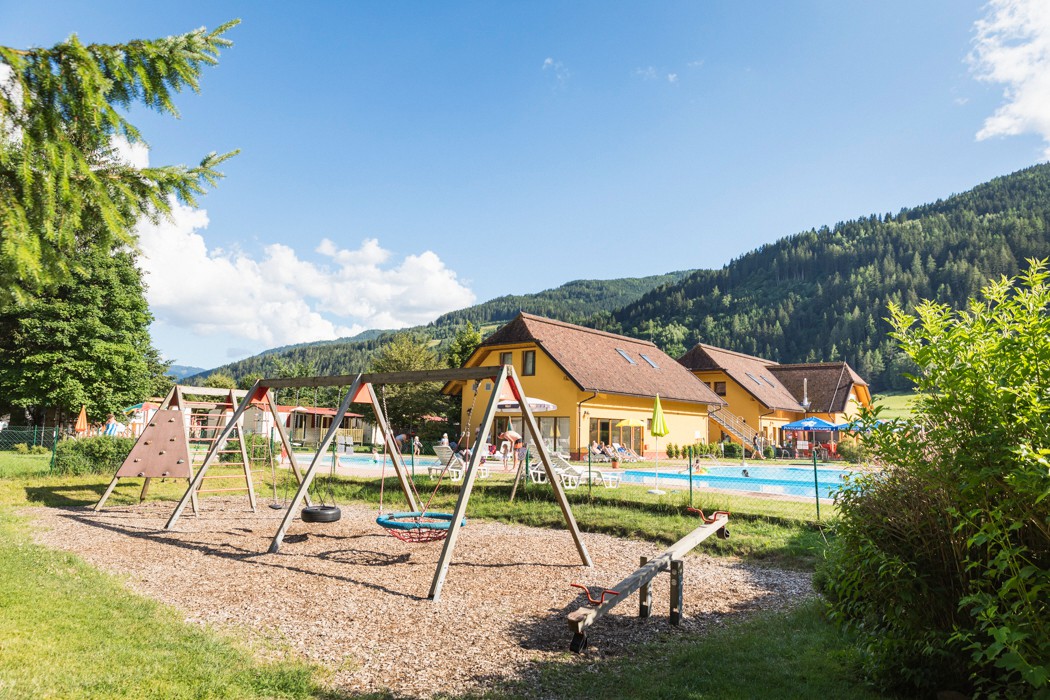 At Bella Austria, playground apparatus featuring swings and a seesaw is positioned in the foreground, adjacent to a swimming pool and yellow structures, encircled by mountains.