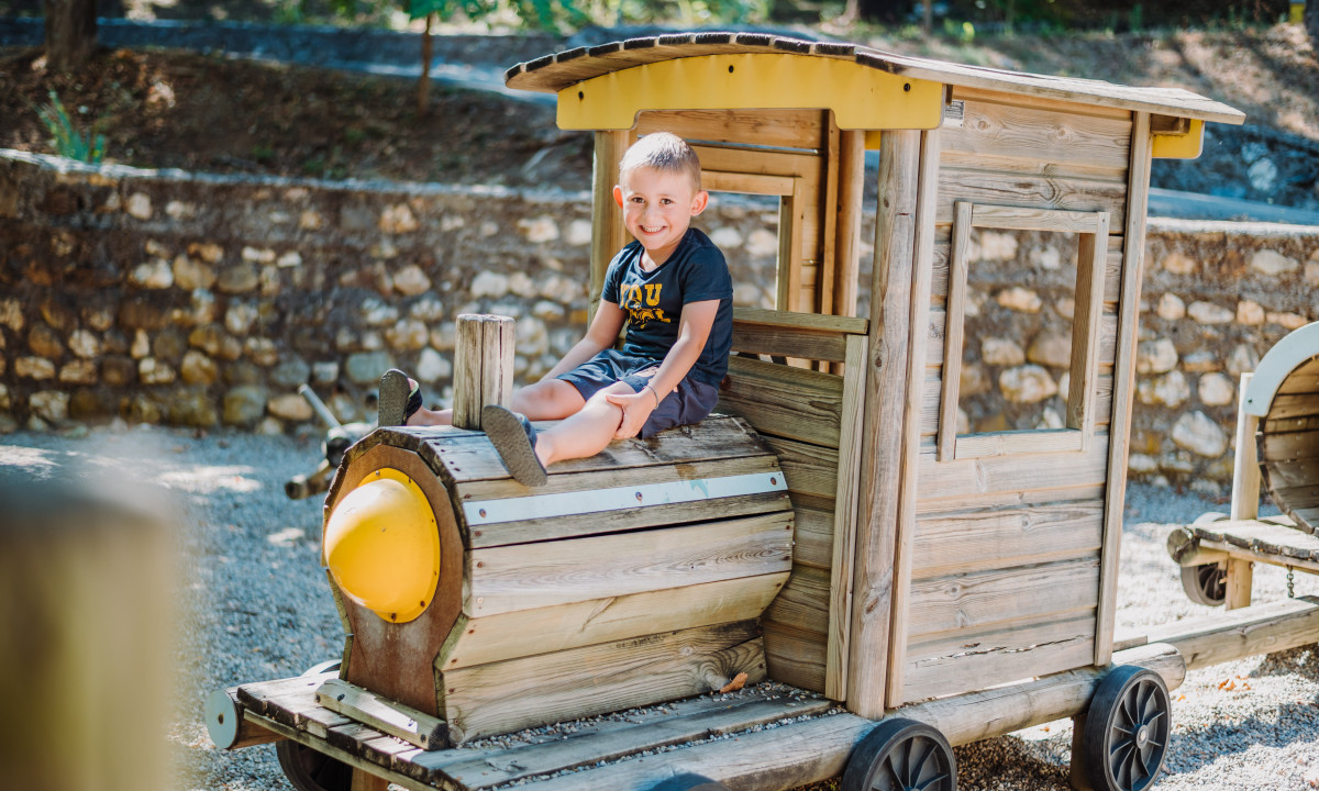 At La Clémentine, a child is perched on a wooden play train, grinning, encircled by trees and a stone wall.