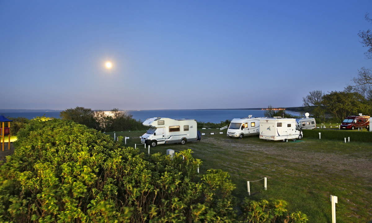 At KNAUS Campingpark Rügen, campers are stationed beneath a bright moon by the ocean, encircled by lush vegetation.