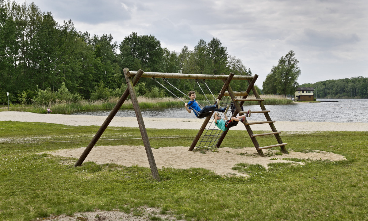 At KNAUS Campingpark Oyten, kids are playing on a wooden play structure next to a lake, encircled by trees and sand.
