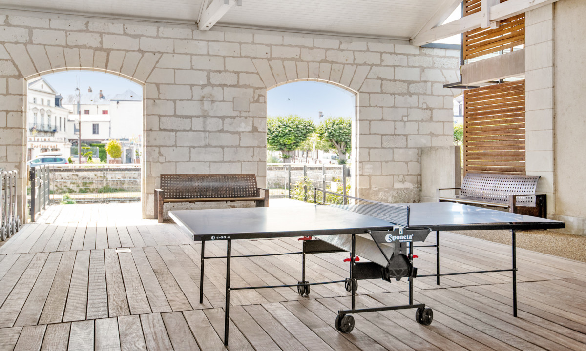 Table tennis table stands on wooden patio beneath an arched stone pavilion, with benches and town buildings visible at Le Moulin des Cordeliers