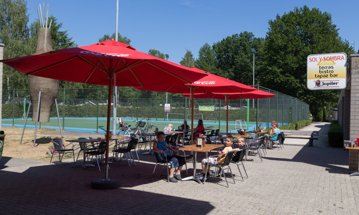 At het Veen, individuals are seated at outdoor café tables beneath crimson umbrellas close to tennis courts, encircled by lush vegetation.