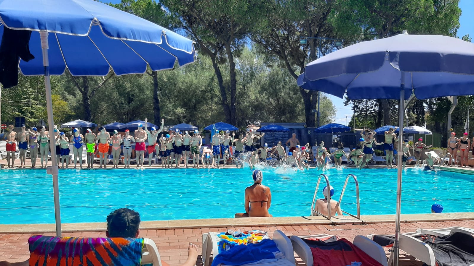 A group jumps into a swimming pool, surrounded by sunbathers and trees under blue umbrellas at Punta Navaccia