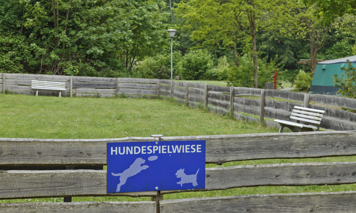 At KNAUS Campingpark Wingst, a blue sign on a wooden fence designates a dog play zone, with white benches and trees encircling it.