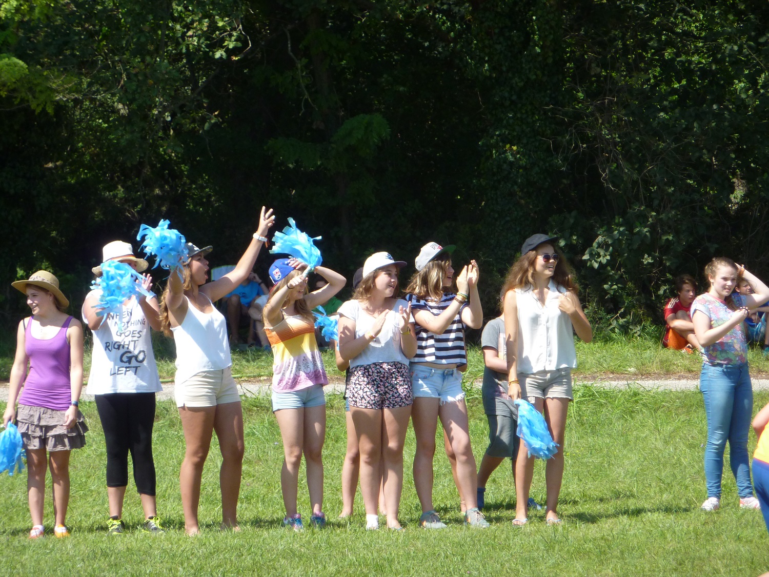A group of people standing, some holding blue pom-poms, gather on a grassy field with trees in the background at Belvedere Pineta