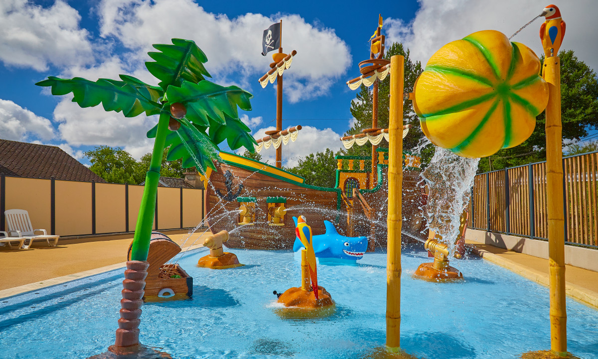 At Terrasses du Lac, there is a pirate-themed aquatic play zone featuring palm trees and a ship, spraying water, enclosed by trees and a fence.