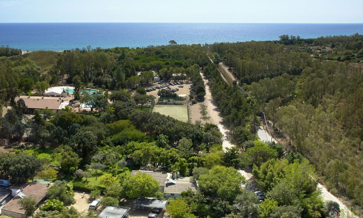 At Ultima Spiaggia, a verdant campsite features structures and vehicles nestled among trees beside a crystal-clear blue sea.