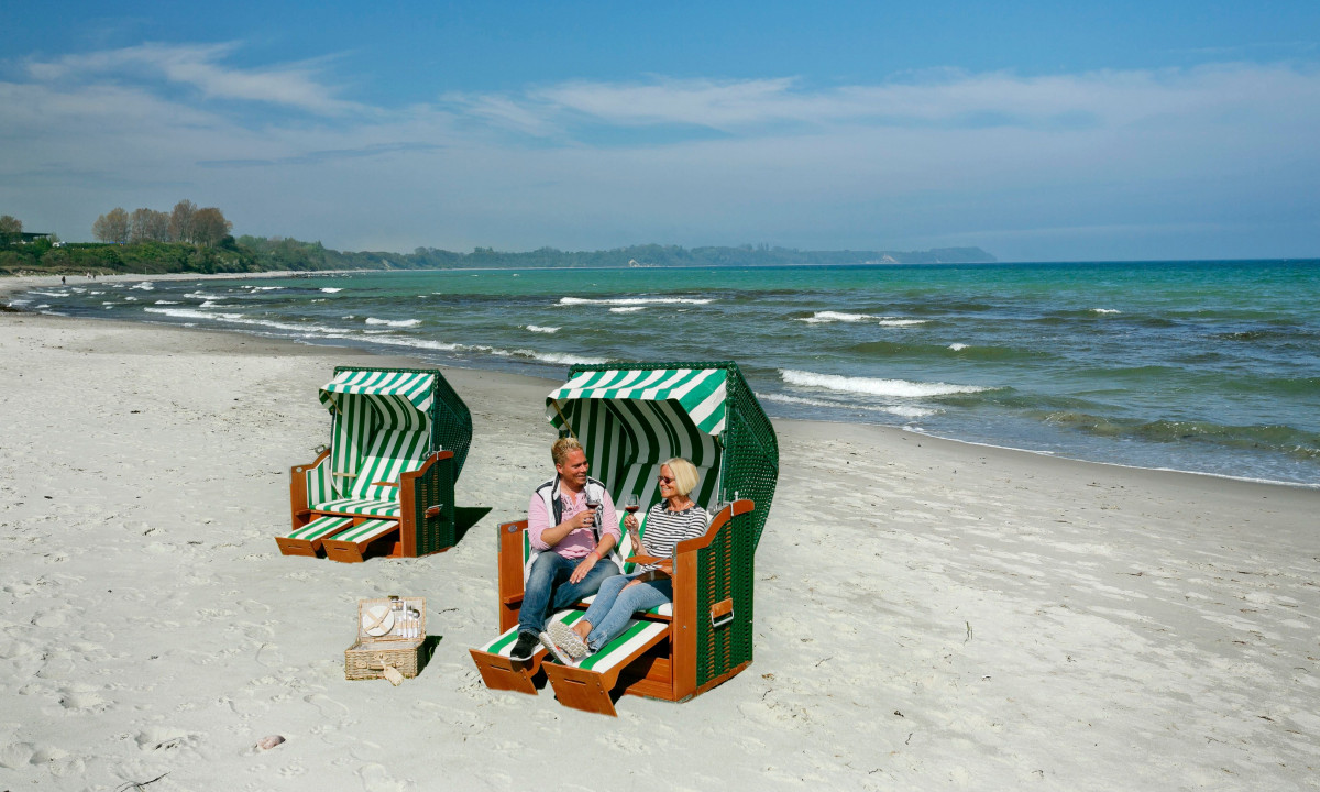 At KNAUS Campingpark Rügen, two individuals are seated in beach chairs, enjoying wine on a sandy shore with the ocean and a distant coastline in view.