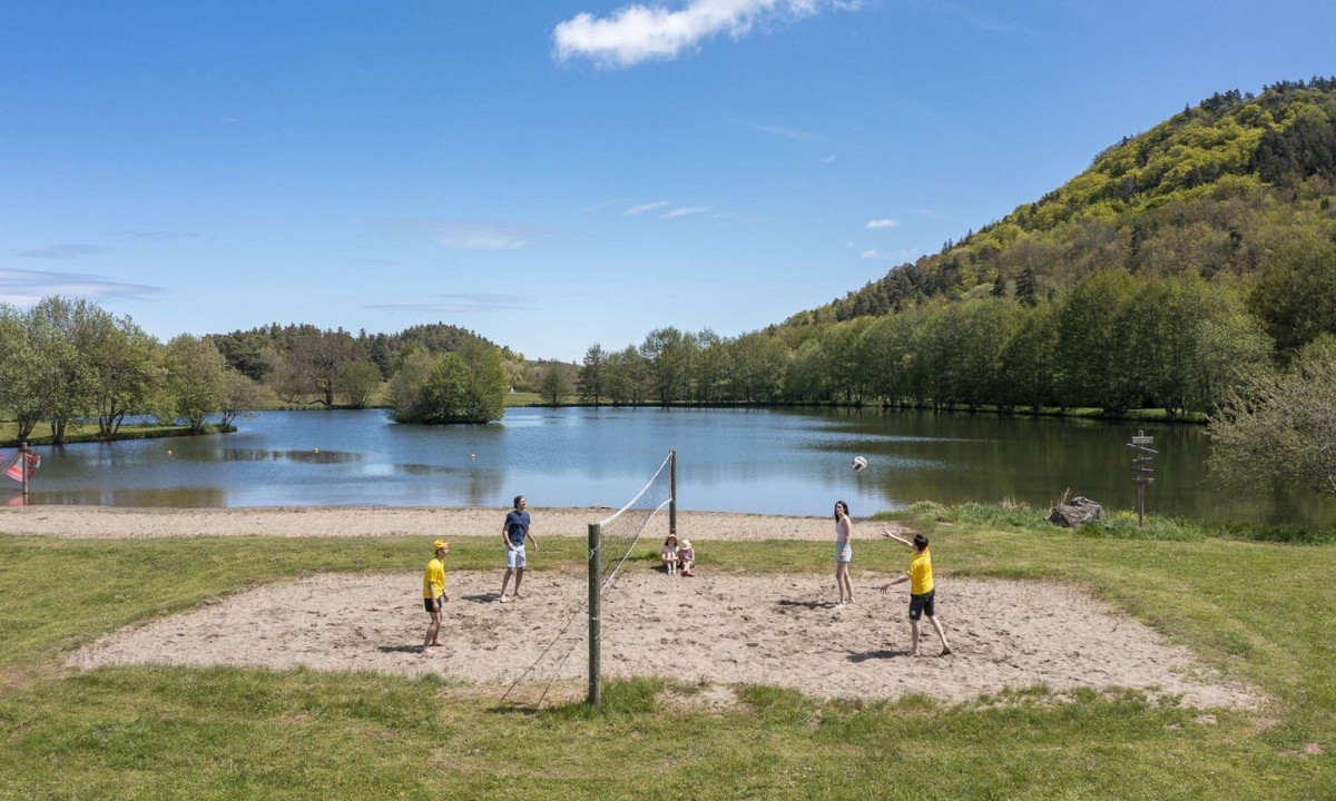 People playing volleyball on a sand court near a lake, surrounded by trees and hills at La Ribeyre