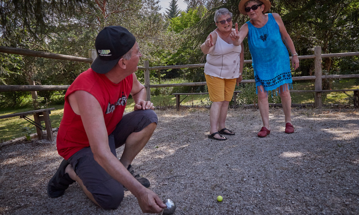 At Le Chêne Vert, three individuals are playing boules, one in a crouched position with balls and two upright, surrounded by trees and a wooden fence.