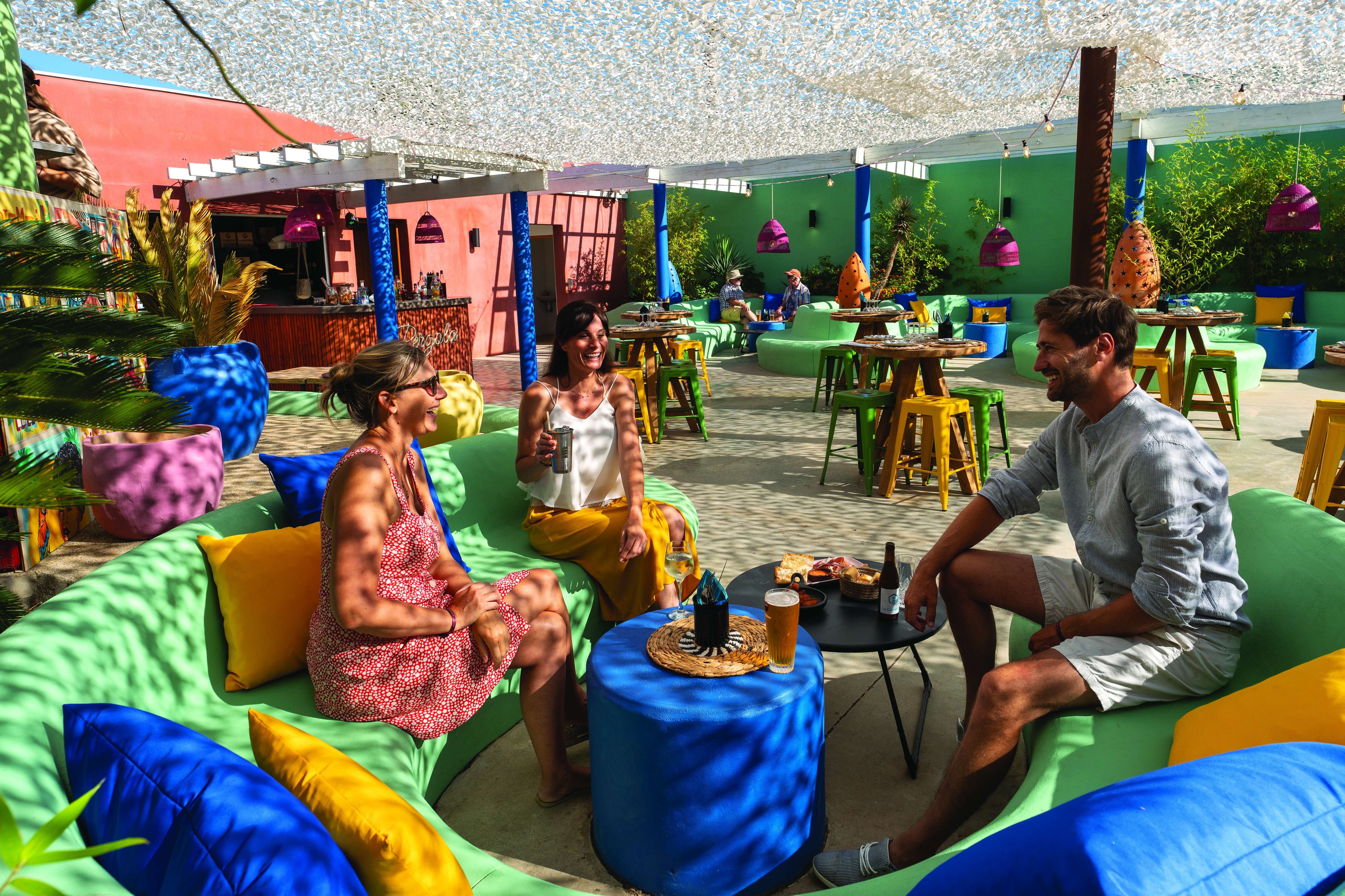 Three people sitting on colorful couches, laughing and enjoying drinks in a sunny, vibrant outdoor lounge at Le Brasilia
