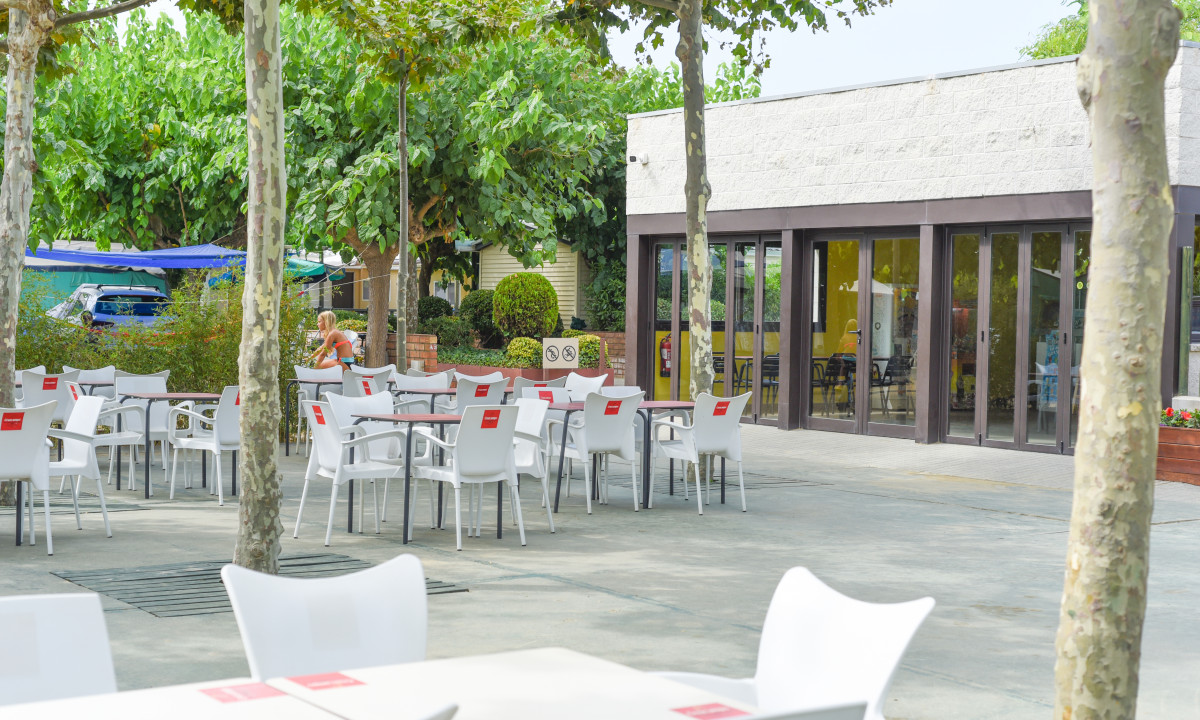 White chairs and tables stand empty under trees beside a modern building at Resort Els Pins