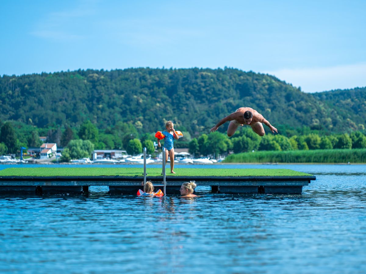 At Lido Verbano, an individual is leaping off a platform into a lake, with children, a hillside, and houses visible in the background.