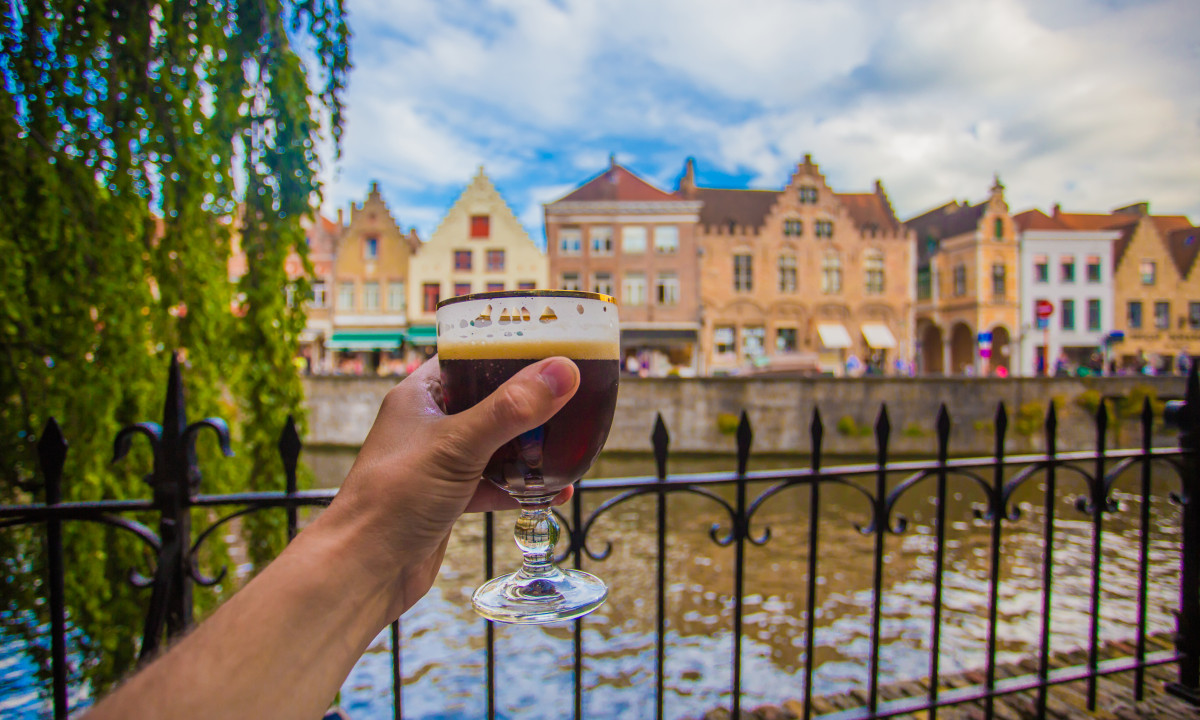 At Urban Gardens Gent, a hand grips a dark brew in a glass, overlooking a charming canal and ancient structures in the backdrop.