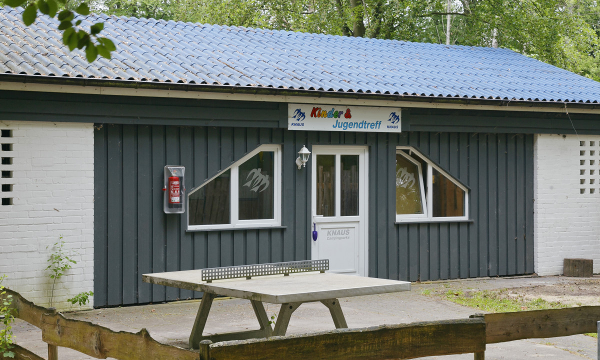 At KNAUS Campingpark Wingst, there is a timber structure featuring a blue rooftop and an outdoor table tennis table, encircled by trees.