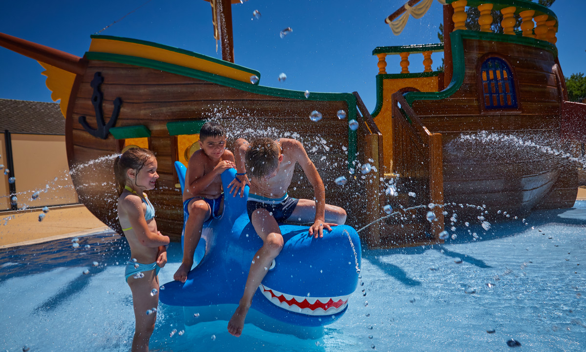 At Terrasses du Lac, three kids are playing on a blue shark-shaped water attraction, close to a wooden pirate vessel in a splash park.