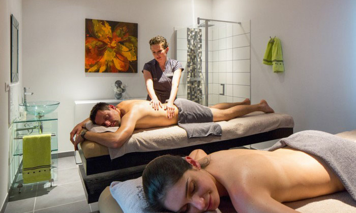 At Les Ranchisses, two individuals are enjoying massages from a therapist in a tranquil, contemporary spa room adorned with artwork and towels.