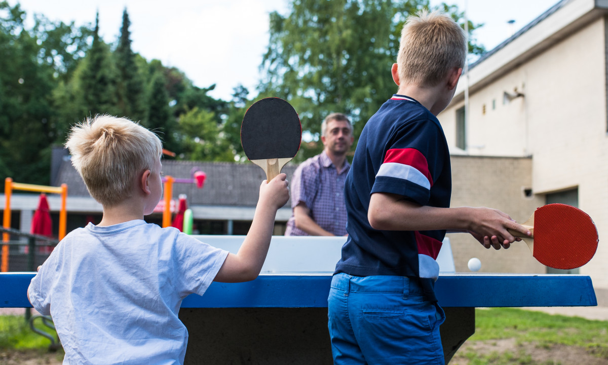 At het Veen, two boys engage in table tennis using paddles and a ball, while an adult observes in a park-like setting.