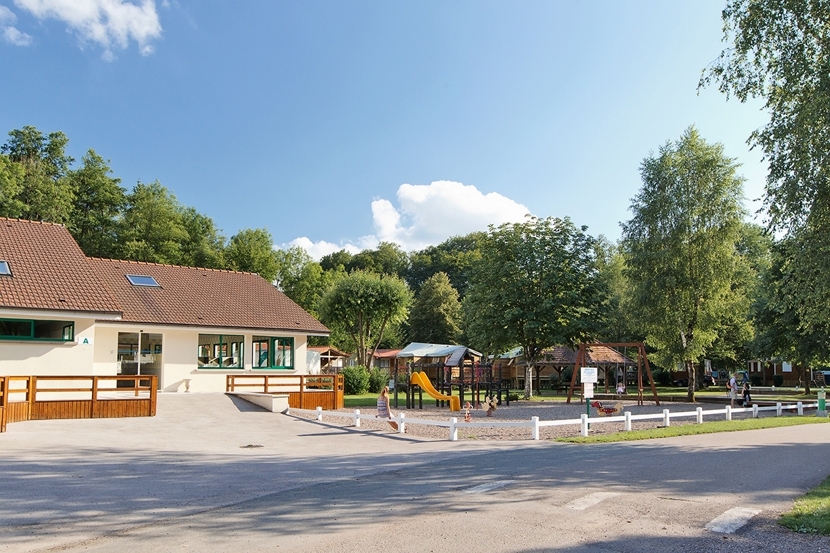 Building adjacent to playground with children playing, surrounded by trees and clear sky at Le Val de Bonnal