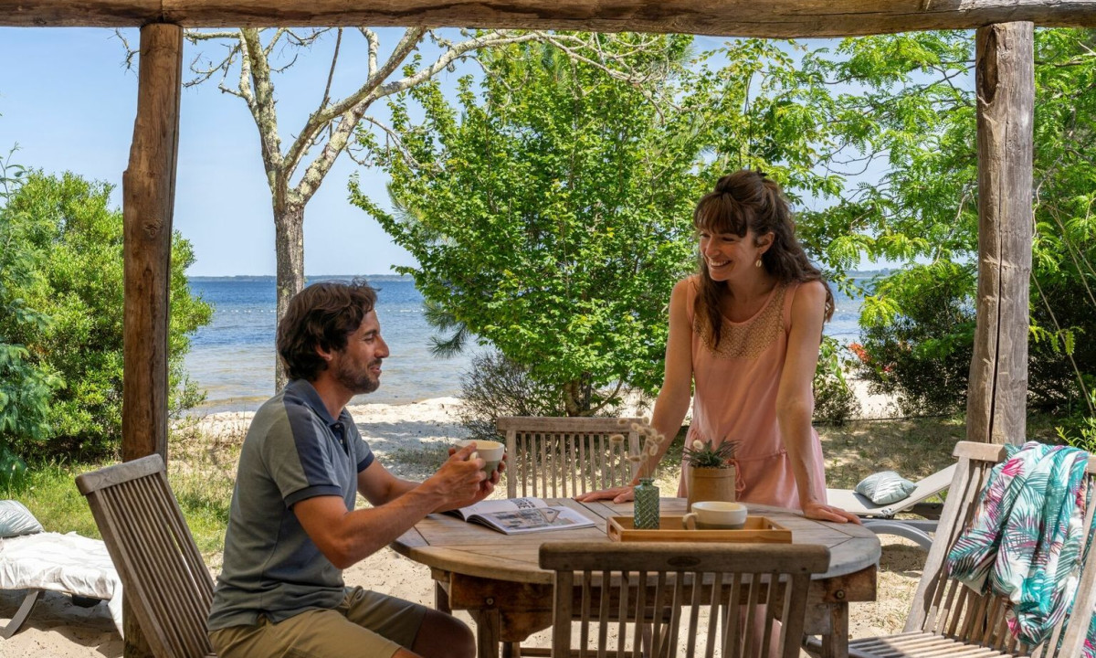 A man and woman converse at a wooden table under a gazebo, with a beach and trees in the background at Maguide