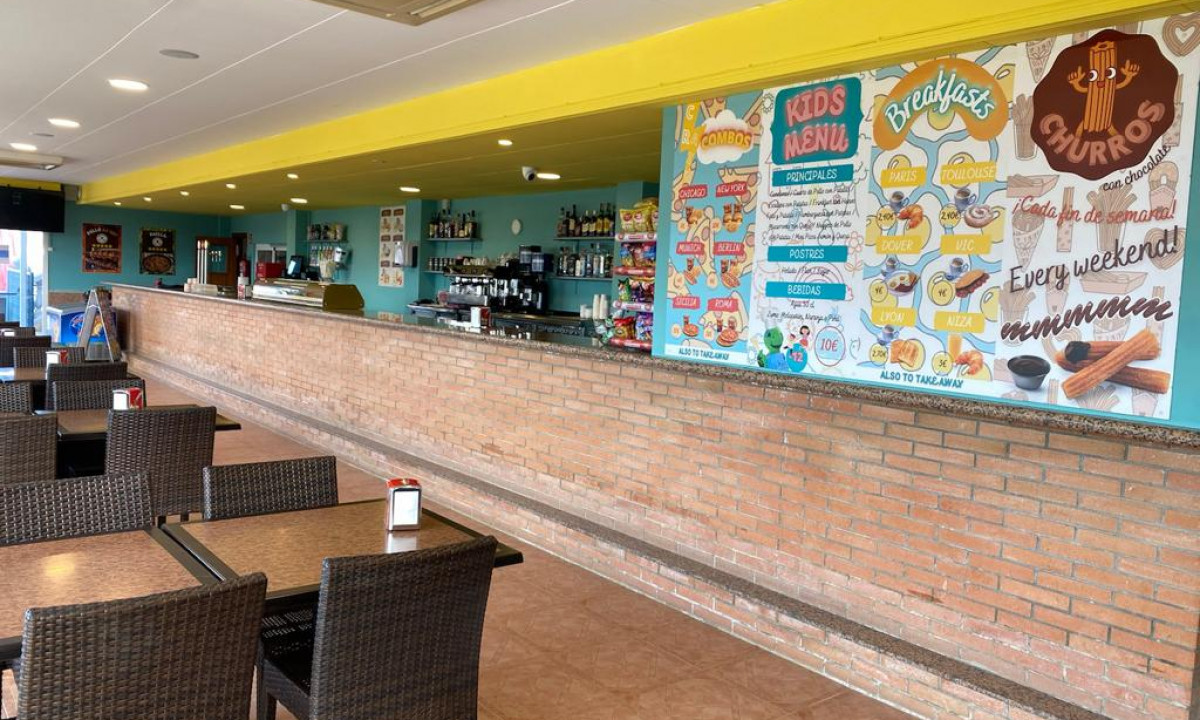 Dining area with wicker chairs and tables, facing a brick bar with colorful menu posters at Resort Els Pins