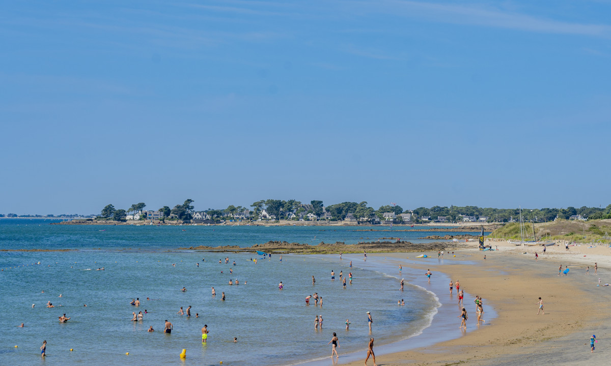 At De la Plage, individuals are swimming and unwinding on a sandy shore beneath a clear azure sky, with residences and foliage in the background.