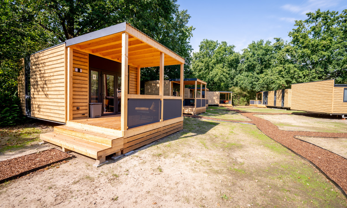 At Urban Gardens Gent, a timber cabin with a veranda is situated in a grassy clearing, encircled by other comparable cabins and trees.