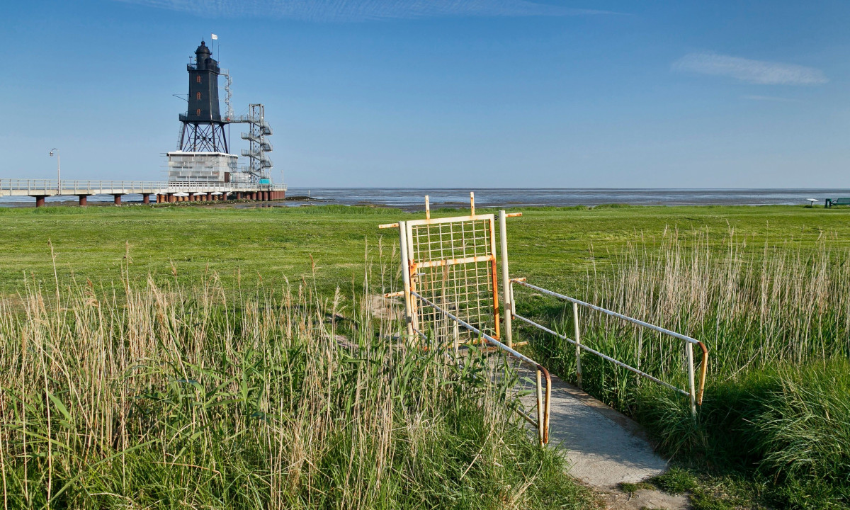 At KNAUS Campingpark Dorum, a beacon by the ocean is accompanied by a grassy meadow and a fenced path in the foreground.