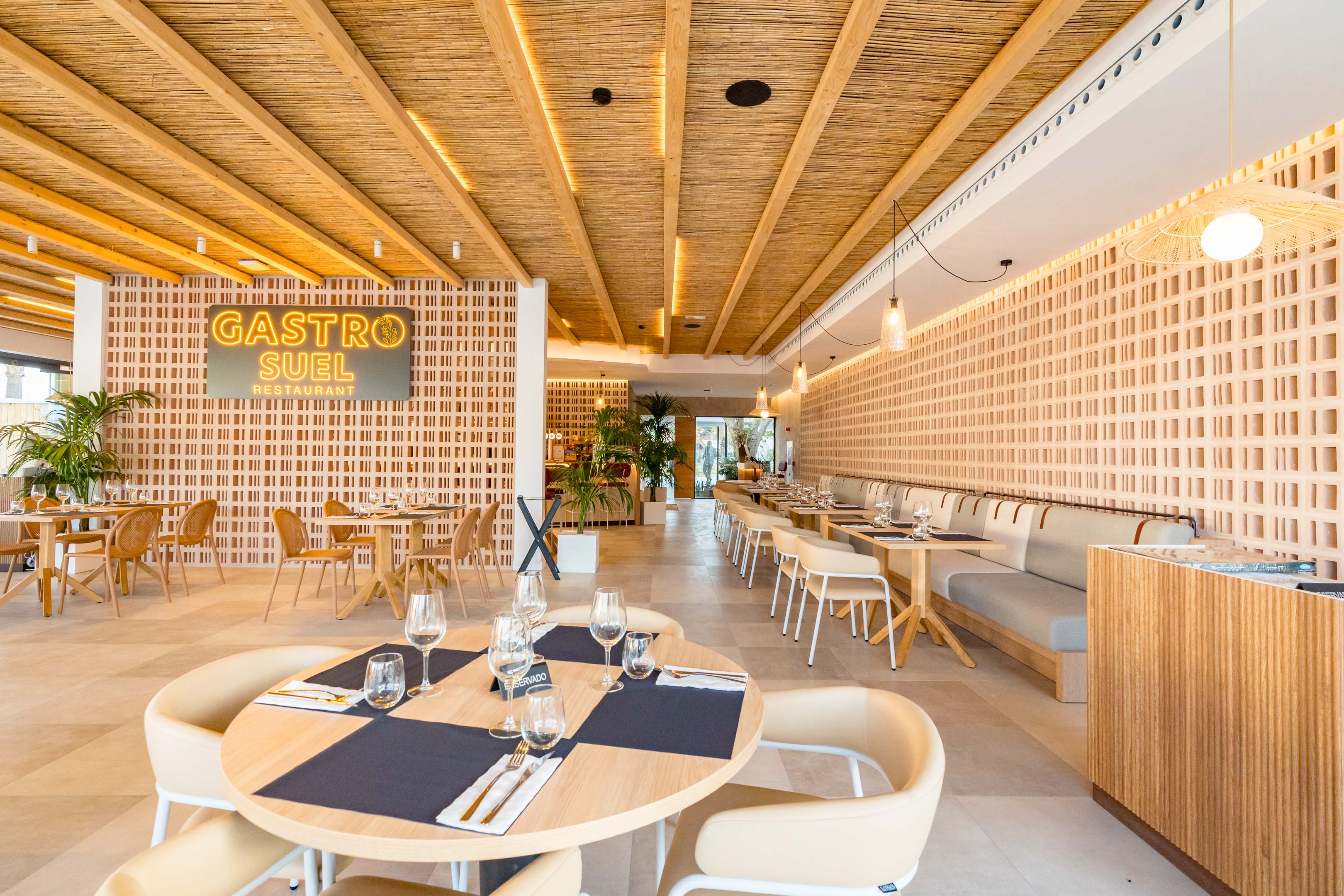 At Costa del Sol Glamping Village, tables are arranged for dining, encircled by light wooden decor and greenery, in a contemporary restaurant atmosphere.