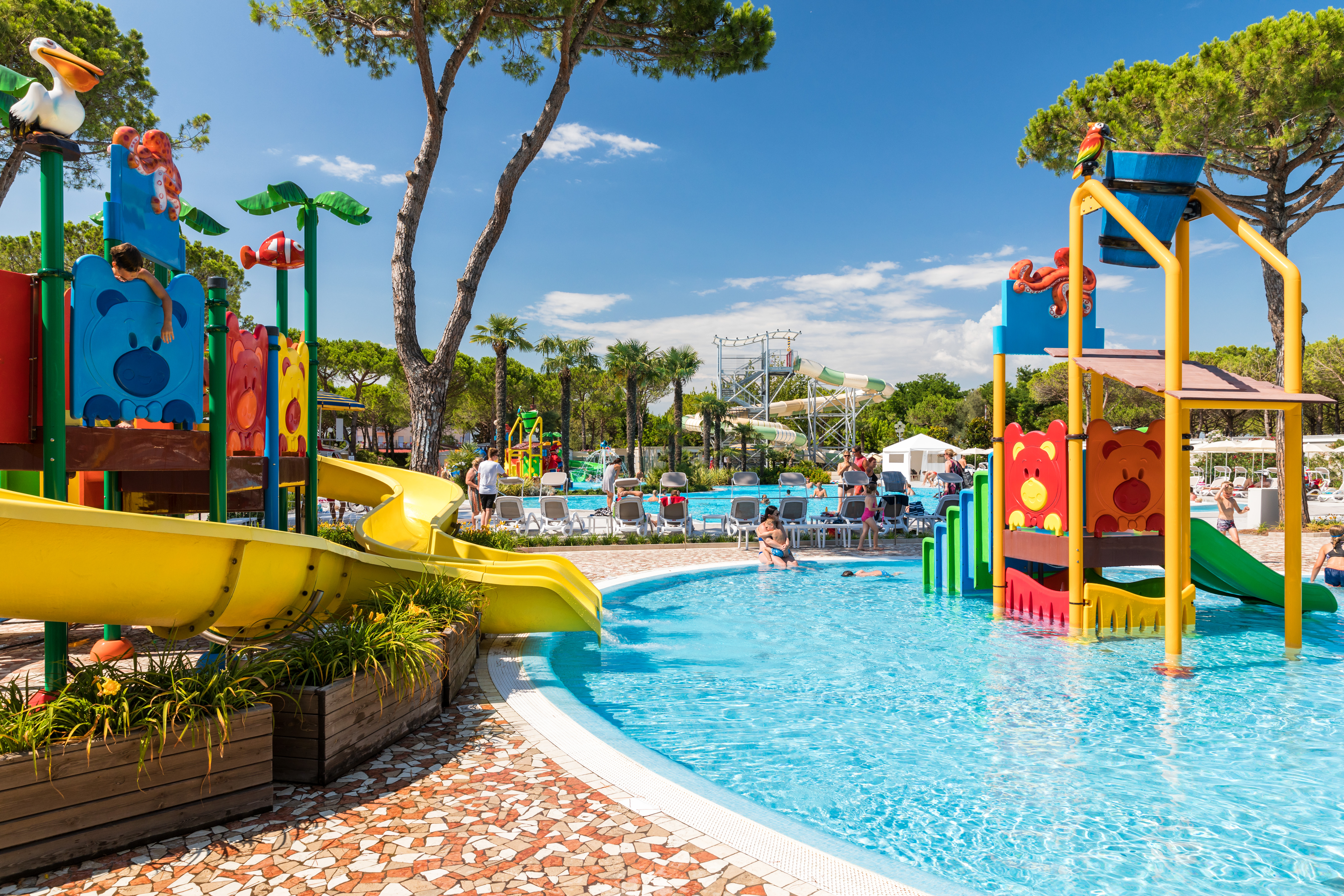 Colorful water playground with slides and climbing structures surrounded by pools and trees, children playing and adults standing nearby at Residence Village