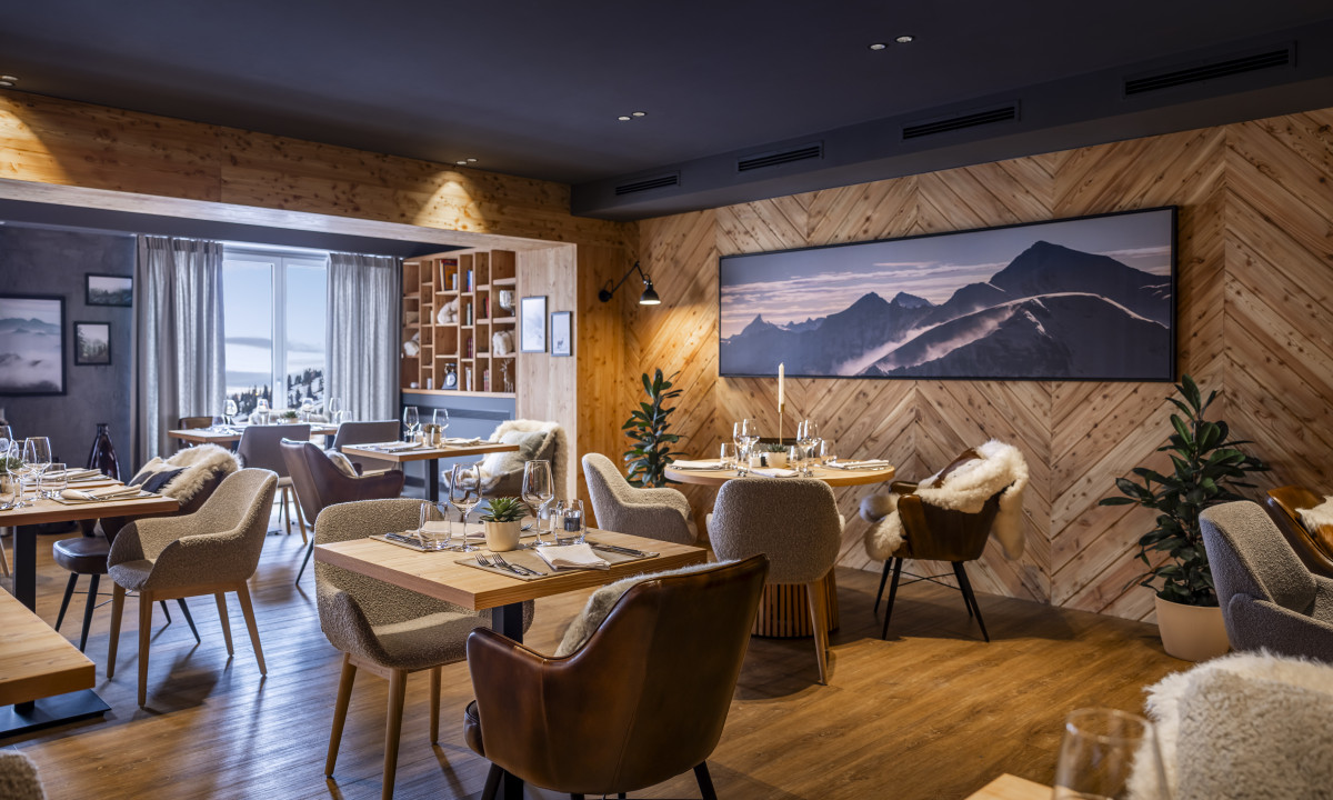 Dining tables set in a cozy restaurant with wooden walls, mountain artwork, and plush chairs; large windows in background at Kesselspitze Valamar Collection Hotel