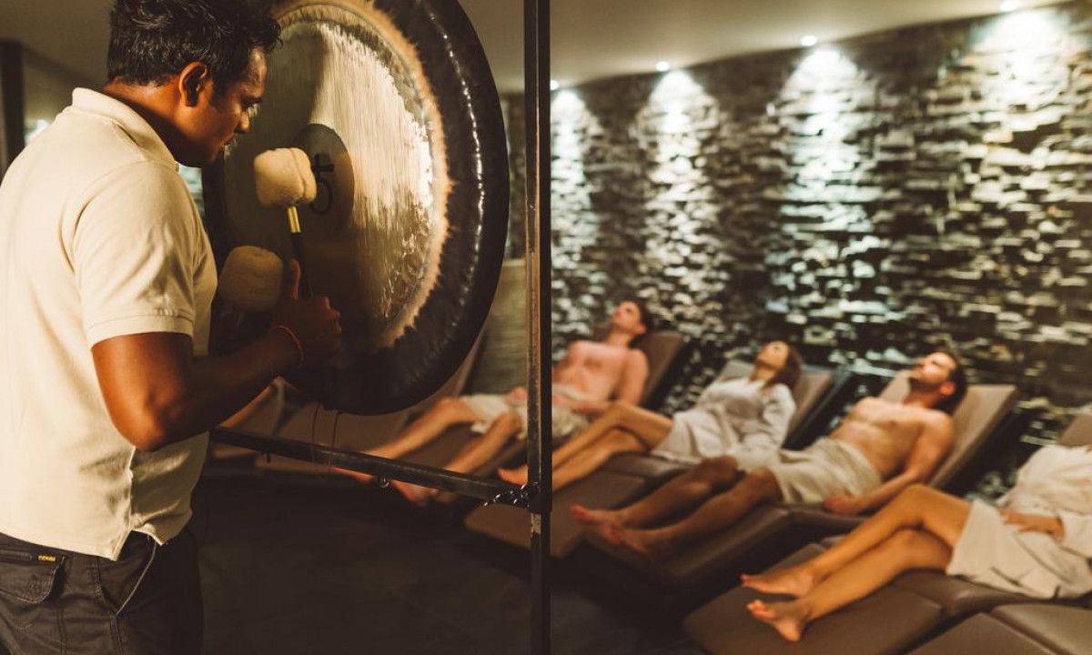 Man striking a gong while four people relax on loungers in a dimly lit room with stone walls at Hotel Sotelia