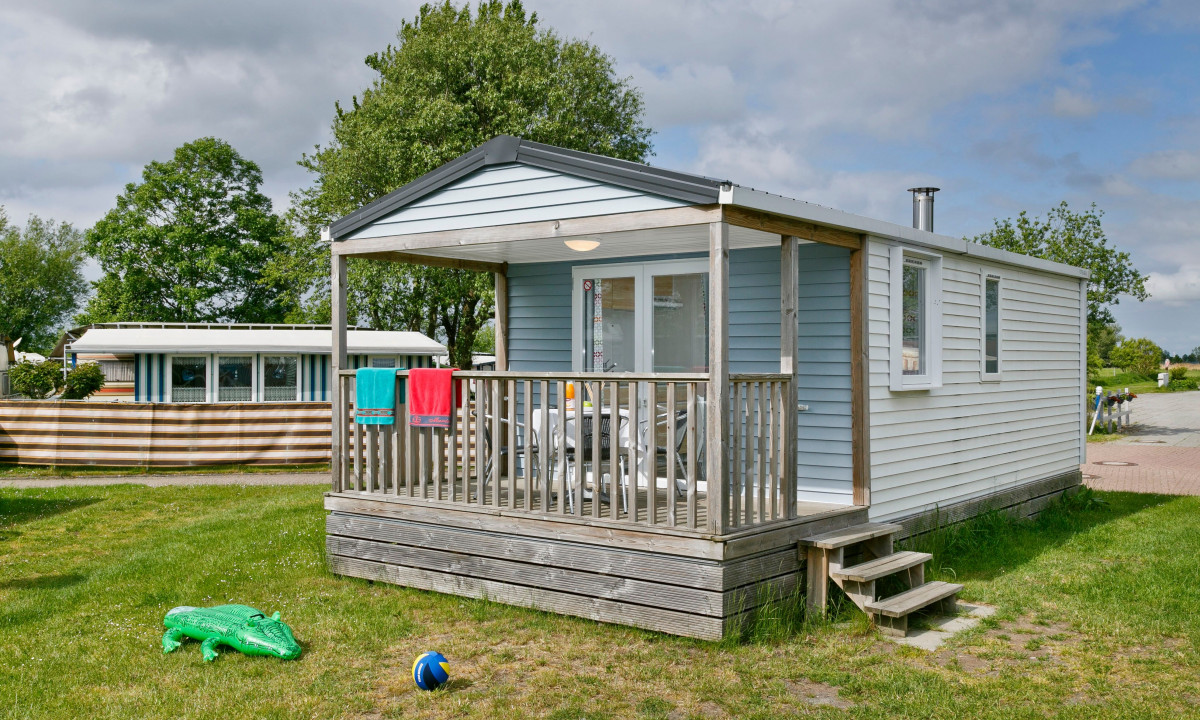 At KNAUS Campingpark Eckwarderhörne, a timber lodge featuring a porch is decorated with hanging towels, encircled by a grassy space that includes an inflatable crocodile and a ball.