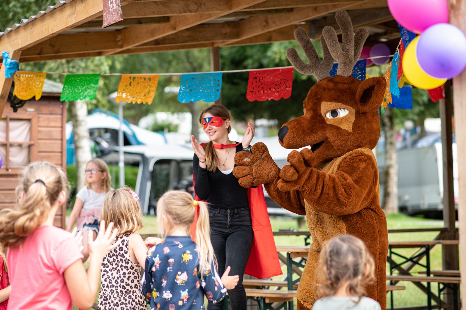 At Petite Suisse, an individual in a crimson mask and reindeer outfit claps, amusing children beneath a wooden shelter.
