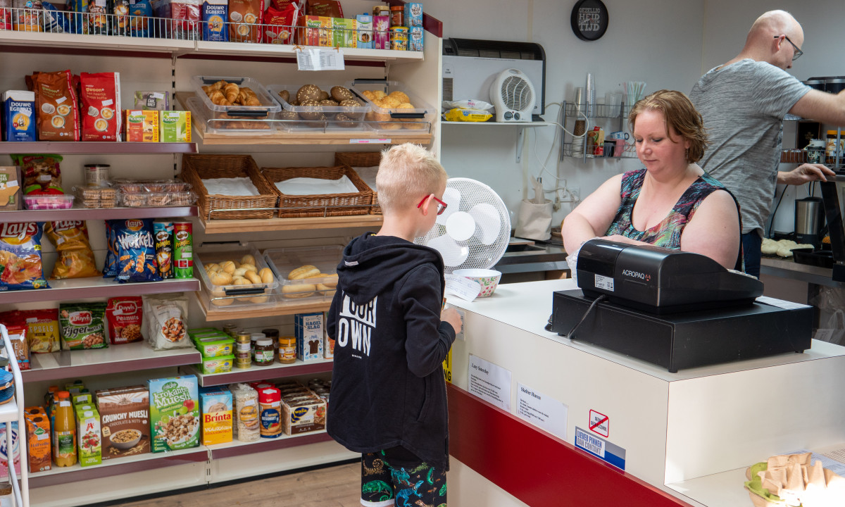 A boy buys items from a cashier at a small, well-stocked snack shop; another person prepares snacks in the background at de Wrange