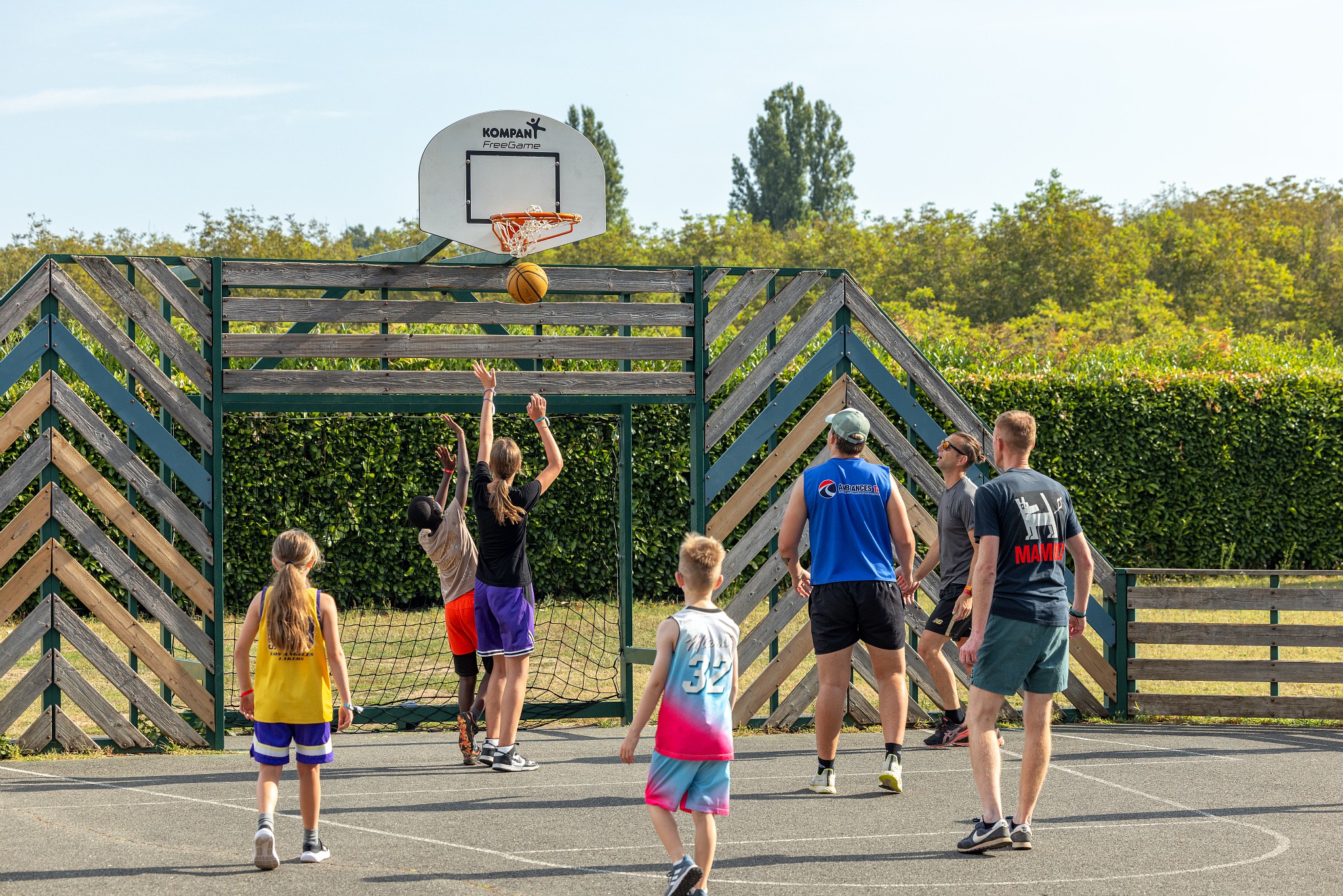 At Marvilla Parks Domaine de Soleil Plage, basketball players are taking shots at a hoop outdoors, encircled by lush greenery, featuring a wooden backboard.