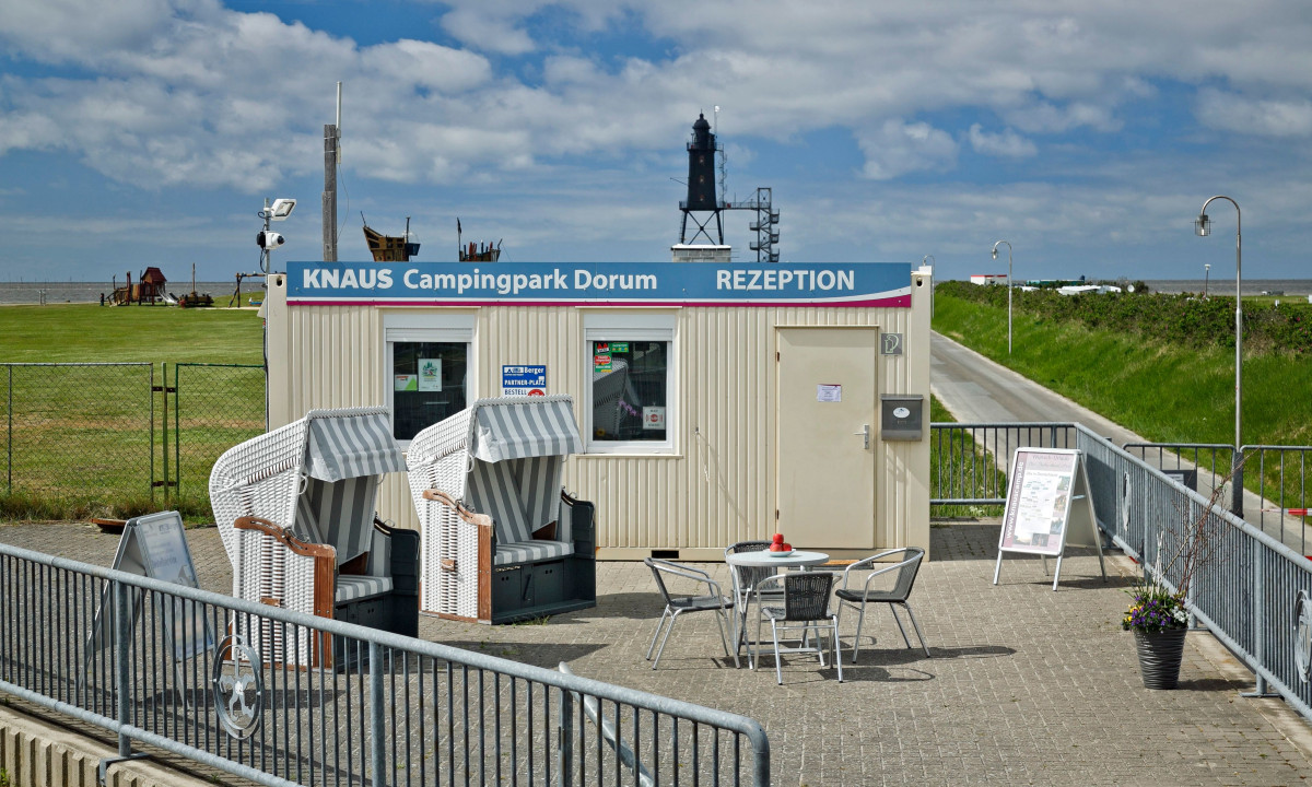 At KNAUS Campingpark Dorum, there is a reception structure featuring a seating area next to lush green fields and a distant lighthouse beneath a cloudy sky.