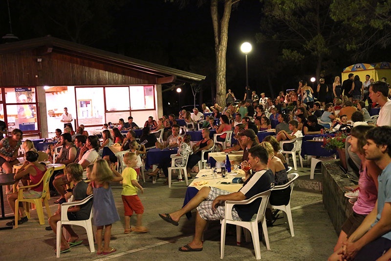 Audience watching a performance, seated outdoors on plastic chairs, with trees and a building in the background at Belvedere Pineta