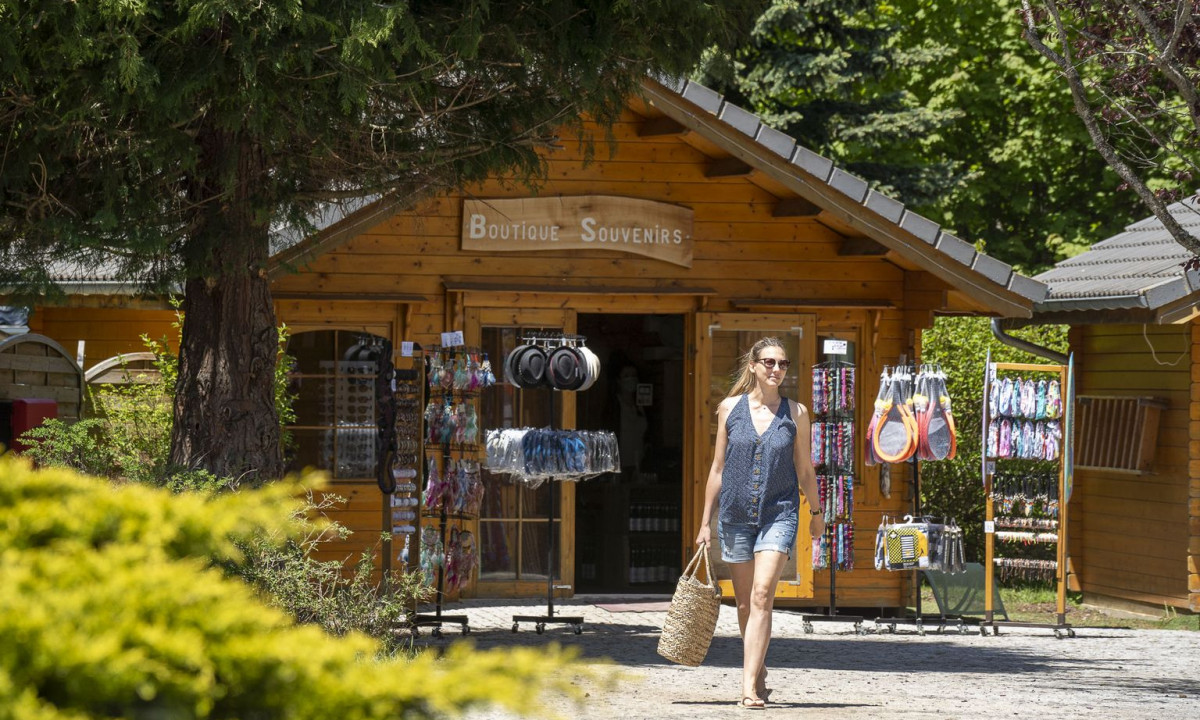 Woman holding basket, walking past souvenir shop with outdoor displays, surrounded by trees and greenery at La Ribeyre