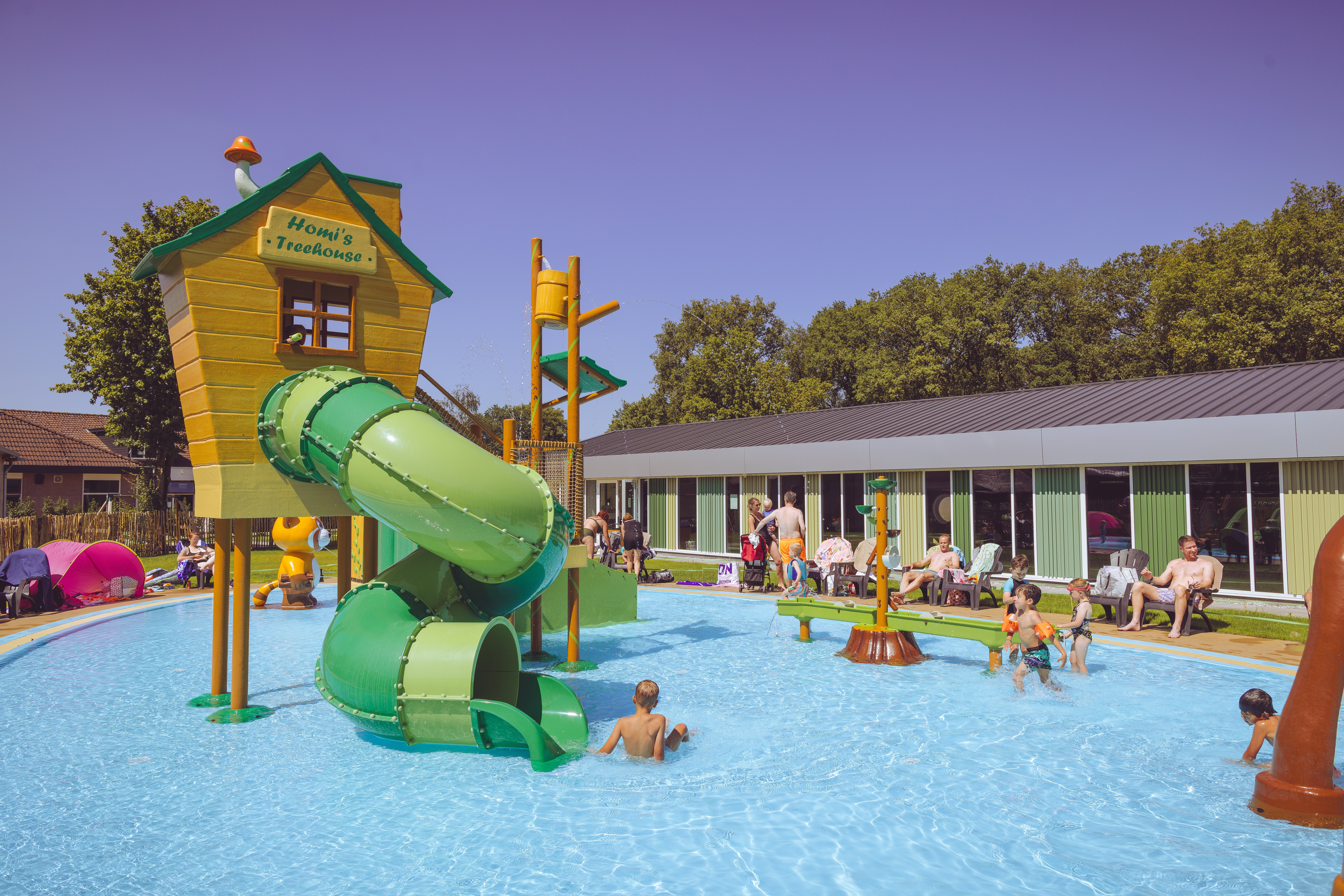 Children playing in a shallow pool with green slides, surrounded by adults and trees at Marvilla Parks Kaatsheuvel